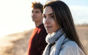 A woman in a gray scarf and coat stands on a beach with a blurred person in the background. The sun illuminates her face, suggesting a chilly day. The background is a sandy coastline under a clear sky.