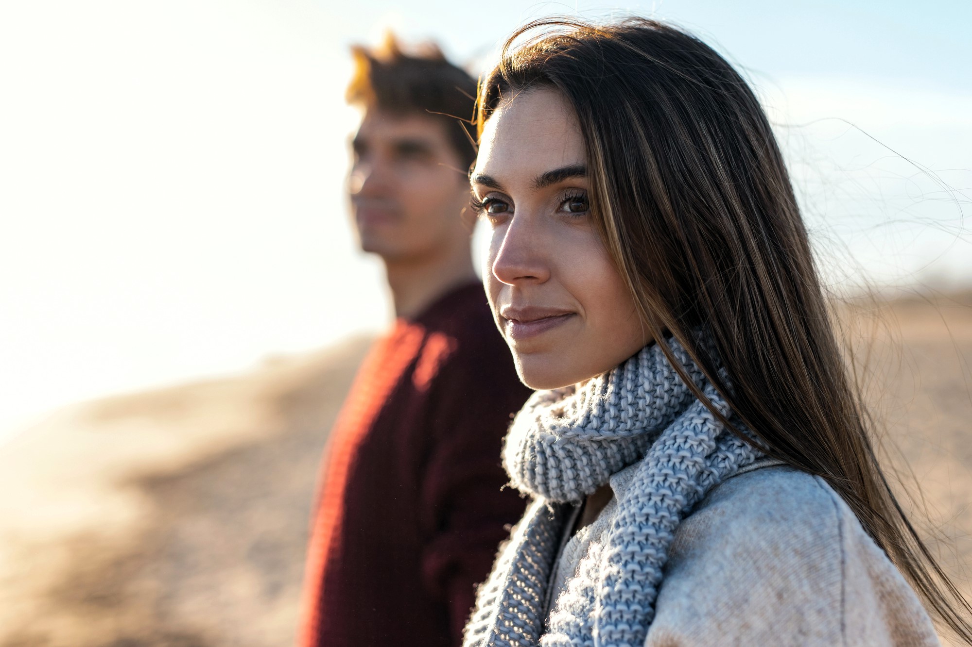 A woman in a gray scarf and coat stands on a beach with a blurred person in the background. The sun illuminates her face, suggesting a chilly day. The background is a sandy coastline under a clear sky.