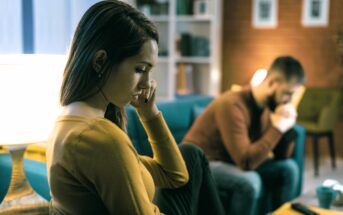 A woman in a yellow sweater sits on a couch, looking thoughtful and touching her face. In the background, a man in a brown sweater sits on another couch, appearing deep in thought. The room is warmly lit with shelves and framed pictures.