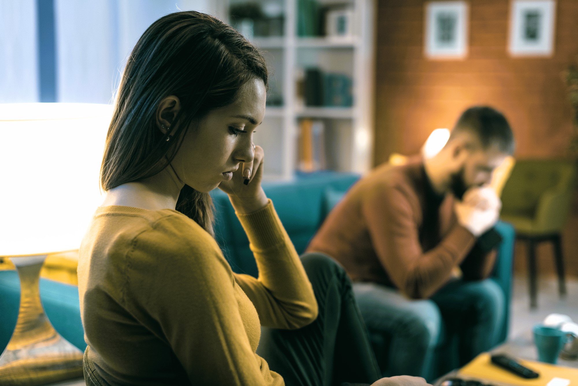 A woman in a yellow sweater sits on a couch, looking thoughtful and touching her face. In the background, a man in a brown sweater sits on another couch, appearing deep in thought. The room is warmly lit with shelves and framed pictures.