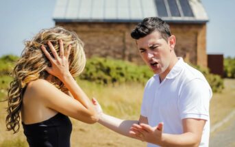 A man and woman appear to be in a heated argument outdoors. The man is gesturing with his hands, looking upset. The woman is holding her head with her hands, looking distressed. They are standing on a dirt path with a building in the background.