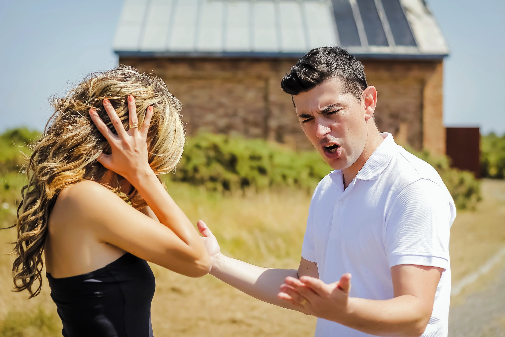 A man and woman appear to be in a heated argument outdoors. The man is gesturing with his hands, looking upset. The woman is holding her head with her hands, looking distressed. They are standing on a dirt path with a building in the background.