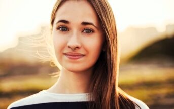 A woman with long brown hair and a septum piercing smiles softly at the camera. She is wearing a black and white top. The background is a blurred outdoor setting with warm, golden light.