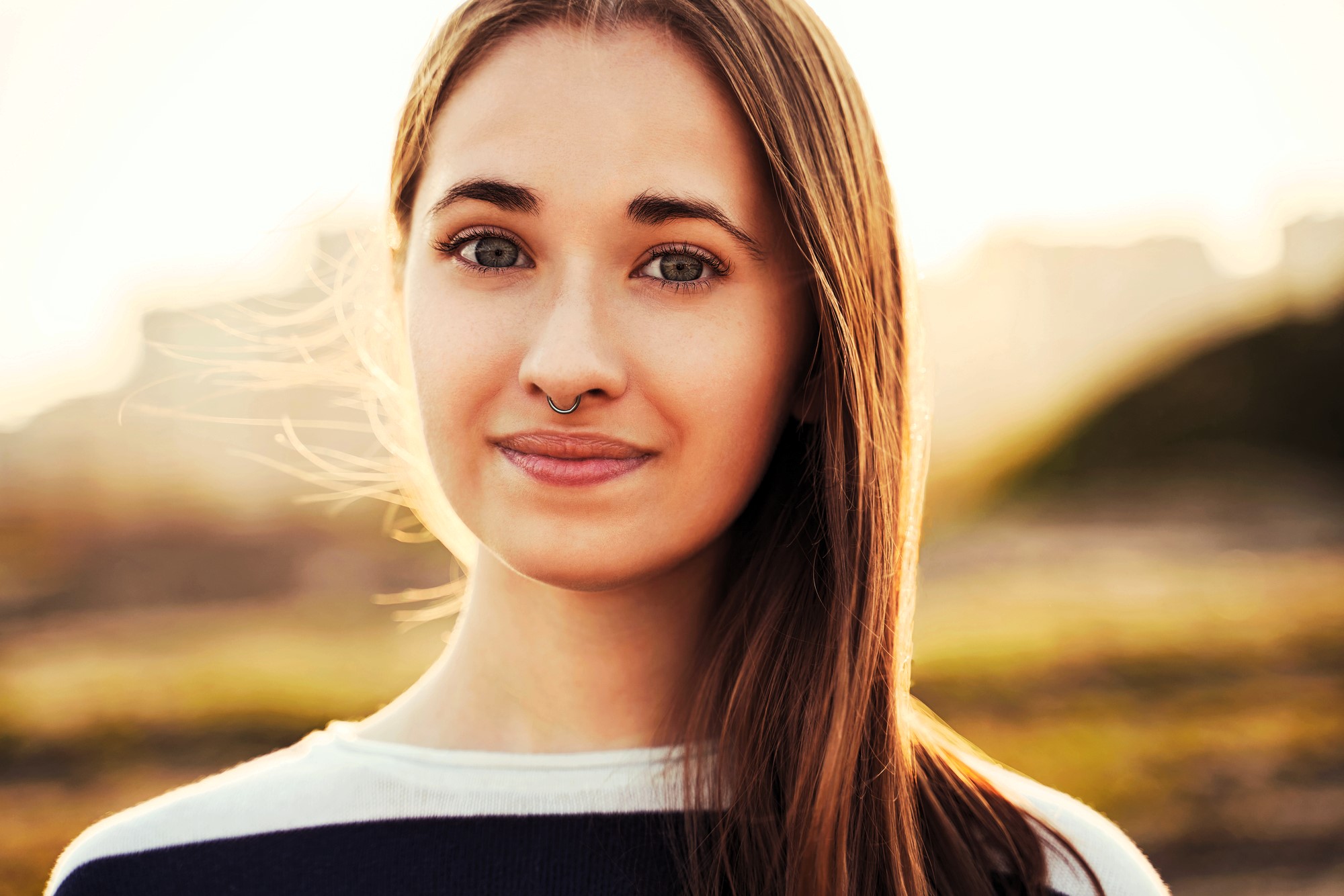 A woman with long brown hair and a septum piercing smiles softly at the camera. She is wearing a black and white top. The background is a blurred outdoor setting with warm, golden light.