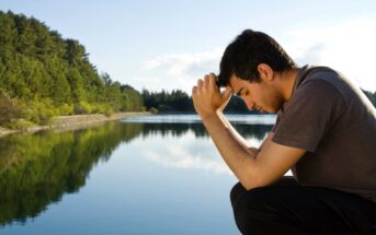 A person crouches near a calm lake, resting their forehead on clasped hands. They appear thoughtful, with a backdrop of green trees under a clear blue sky, reflecting on the water's surface.