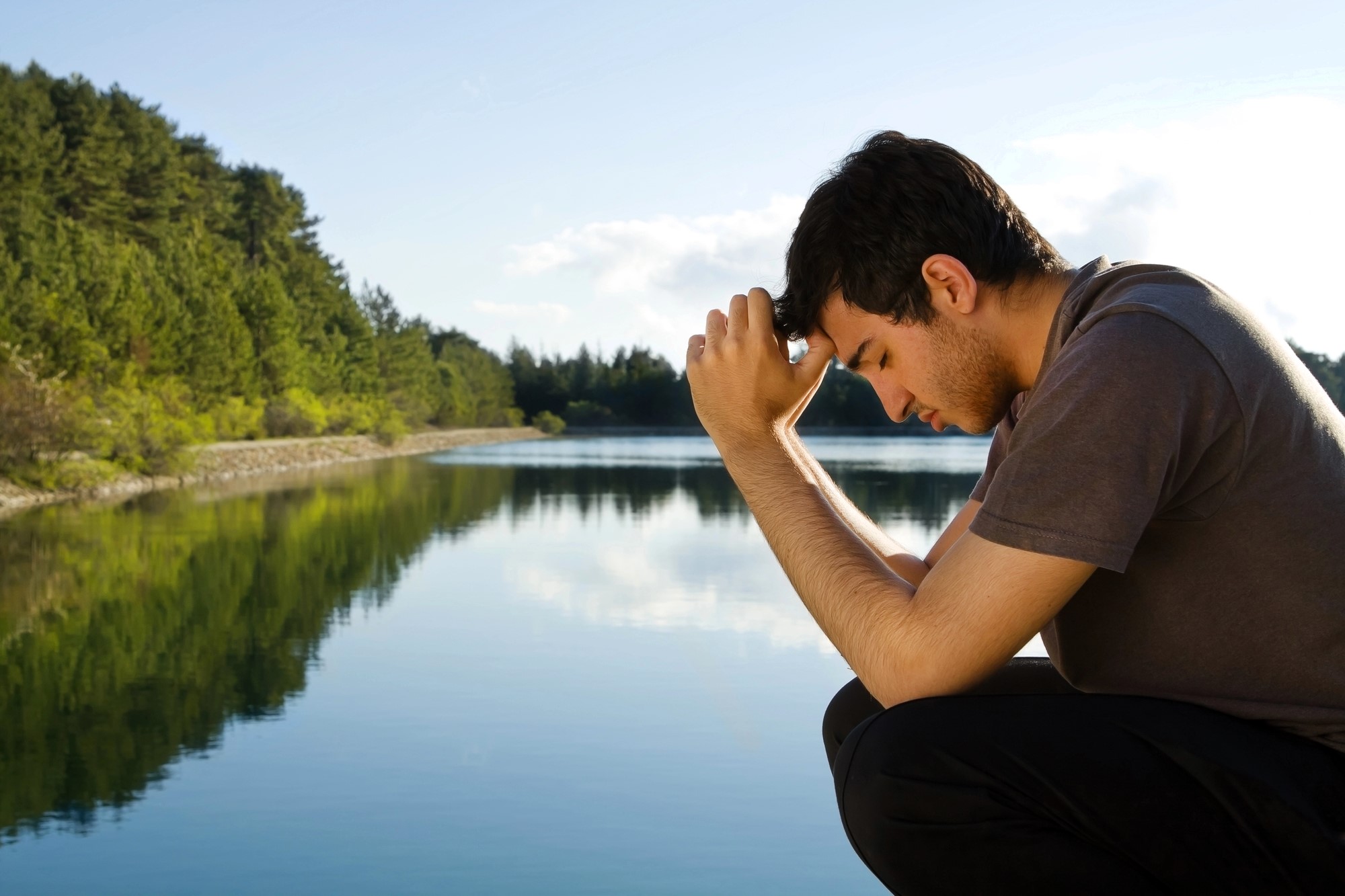 A person crouches near a calm lake, resting their forehead on clasped hands. They appear thoughtful, with a backdrop of green trees under a clear blue sky, reflecting on the water's surface.