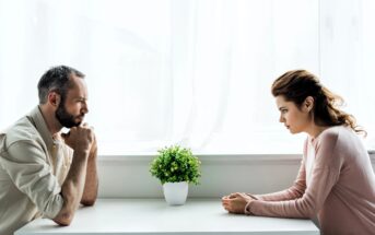 A man and a woman sit across from each other at a white table, with a small potted plant between them. Both appear deep in thought or conversation, in a brightly lit room with sheer white curtains.