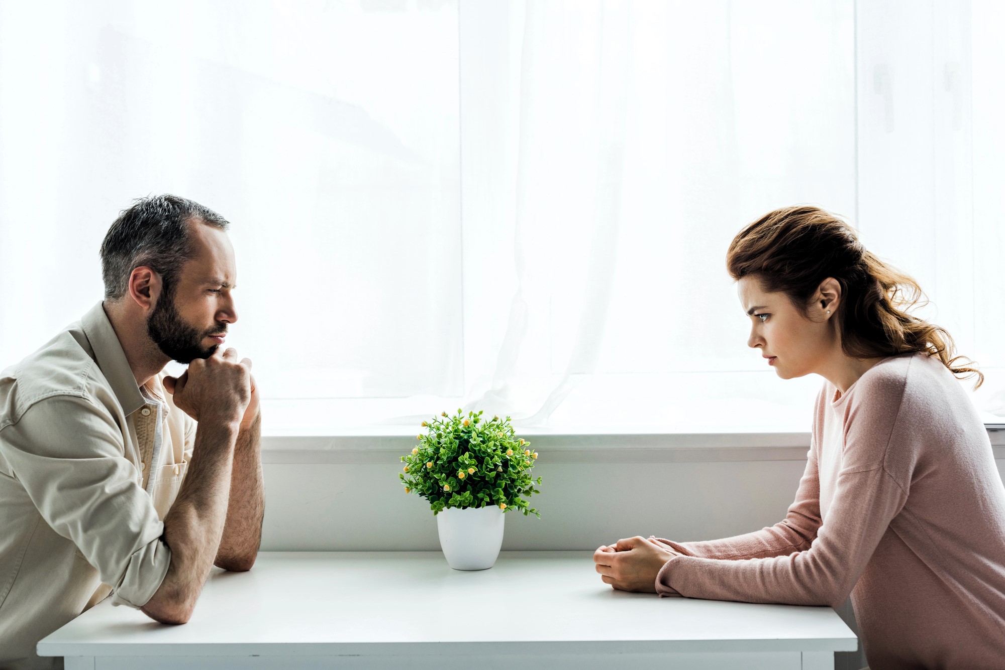 A man and a woman sit across from each other at a white table, with a small potted plant between them. Both appear deep in thought or conversation, in a brightly lit room with sheer white curtains.