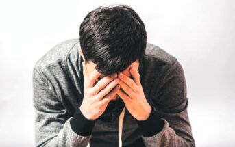 A person with short dark hair, wearing a gray hoodie, sits with their hands covering their face in a gesture of distress or contemplation against a plain background.