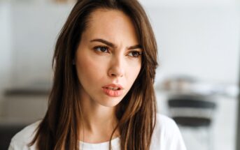 A woman with long brown hair looks thoughtfully to the side. She wears a white shirt, and the background is blurred with indistinct furniture, conveying a focused and contemplative mood.