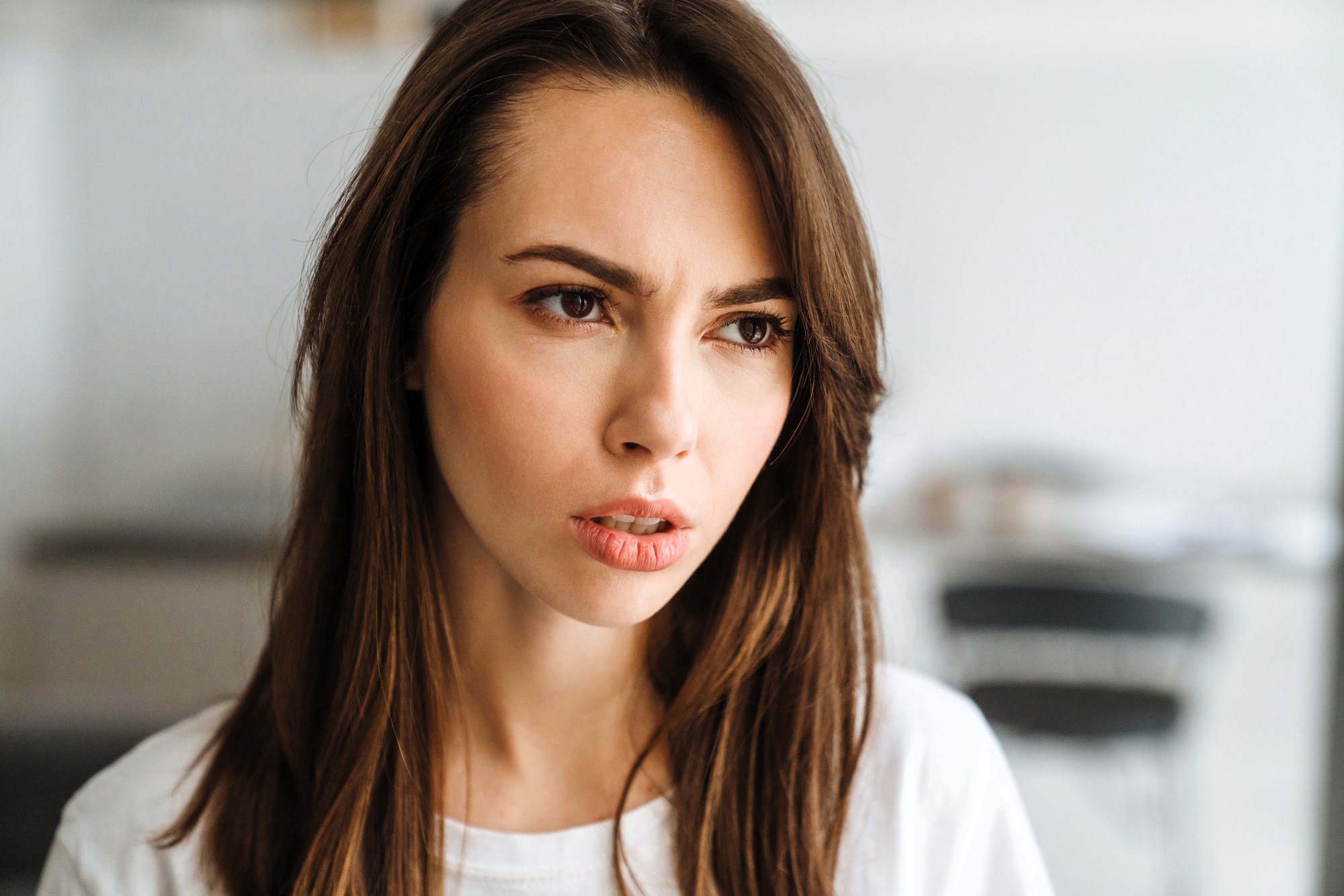 A woman with long brown hair looks thoughtfully to the side. She wears a white shirt, and the background is blurred with indistinct furniture, conveying a focused and contemplative mood.