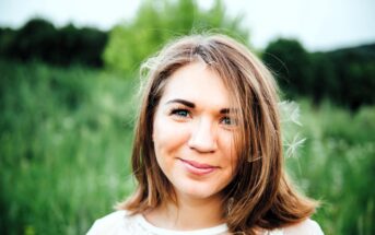 A woman with brown hair smiles softly at the camera, standing in a lush green field with trees in the background. She wears a light-colored, semi-transparent top, and a few strands of her hair are blown by the wind.