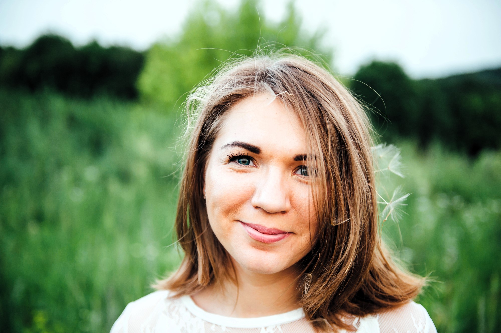 A woman with brown hair smiles softly at the camera, standing in a lush green field with trees in the background. She wears a light-colored, semi-transparent top, and a few strands of her hair are blown by the wind.