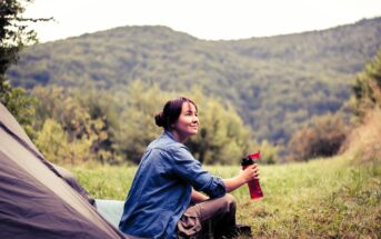 A woman in a blue shirt sits on the grass near a tent, holding a red water bottle. She looks content, surrounded by a lush green landscape with hills in the background. The scene conveys a peaceful outdoor camping experience.