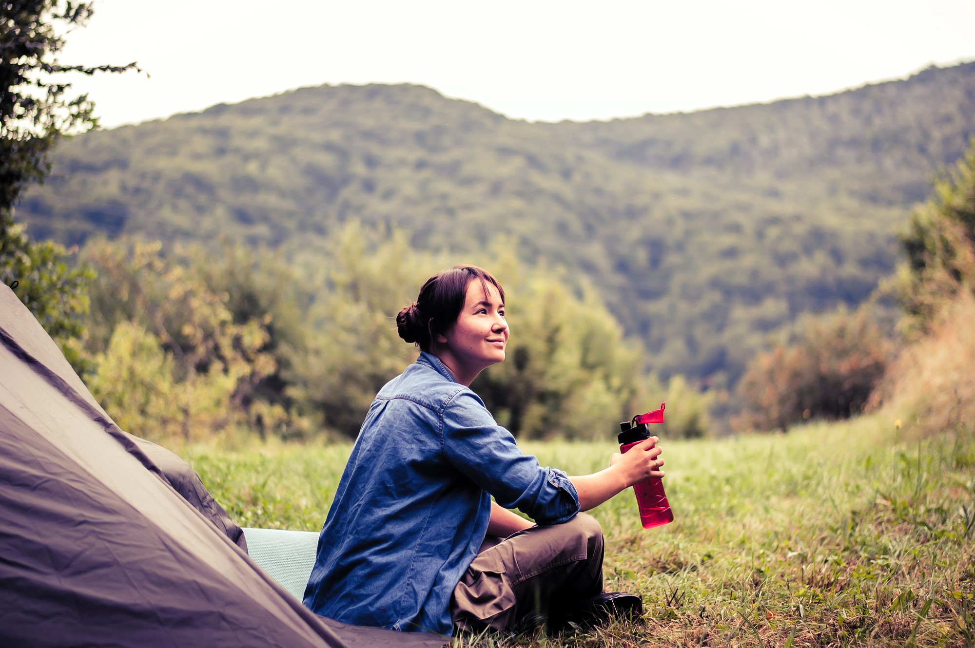 A woman in a blue shirt sits on the grass near a tent, holding a red water bottle. She looks content, surrounded by a lush green landscape with hills in the background. The scene conveys a peaceful outdoor camping experience.