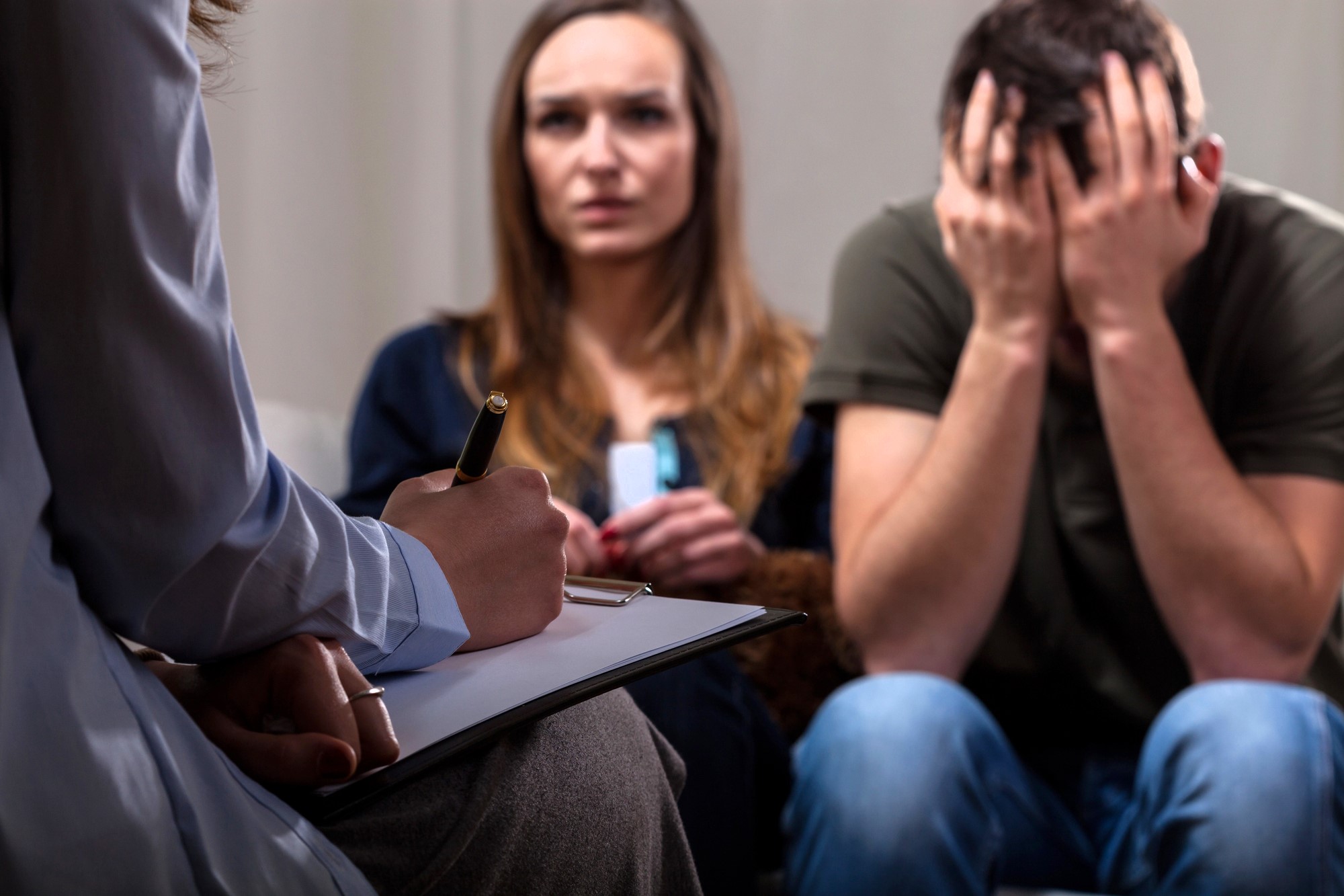 A person is sitting on a chair holding a clipboard and pen, taking notes. In the background, a woman sits looking concerned and a man beside her has his head in his hands, appearing distressed.