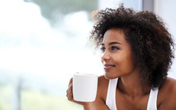 A woman with curly hair smiles gently while holding a white mug. She is sitting by a window, and the background is softly blurred, giving a serene and relaxed atmosphere.