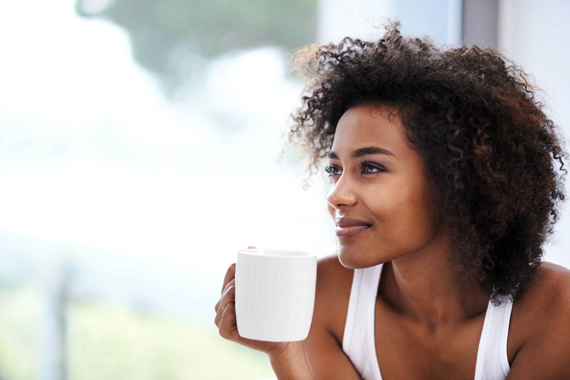 A woman with curly hair smiles gently while holding a white mug. She is sitting by a window, and the background is softly blurred, giving a serene and relaxed atmosphere.