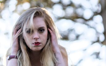 A woman with long blonde hair and red lipstick looks intently at the camera. She holds her hands to her head, with an out-of-focus background of trees and sunlight.