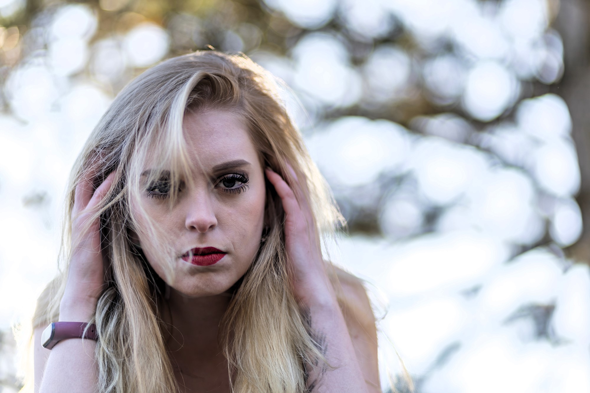 A woman with long blonde hair and red lipstick looks intently at the camera. She holds her hands to her head, with an out-of-focus background of trees and sunlight.