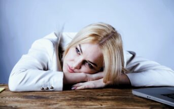 A woman with blonde hair, wearing a white blouse, rests her head on her folded arms on a wooden table. She appears thoughtful or tired, with a laptop partially visible nearby against a plain background.