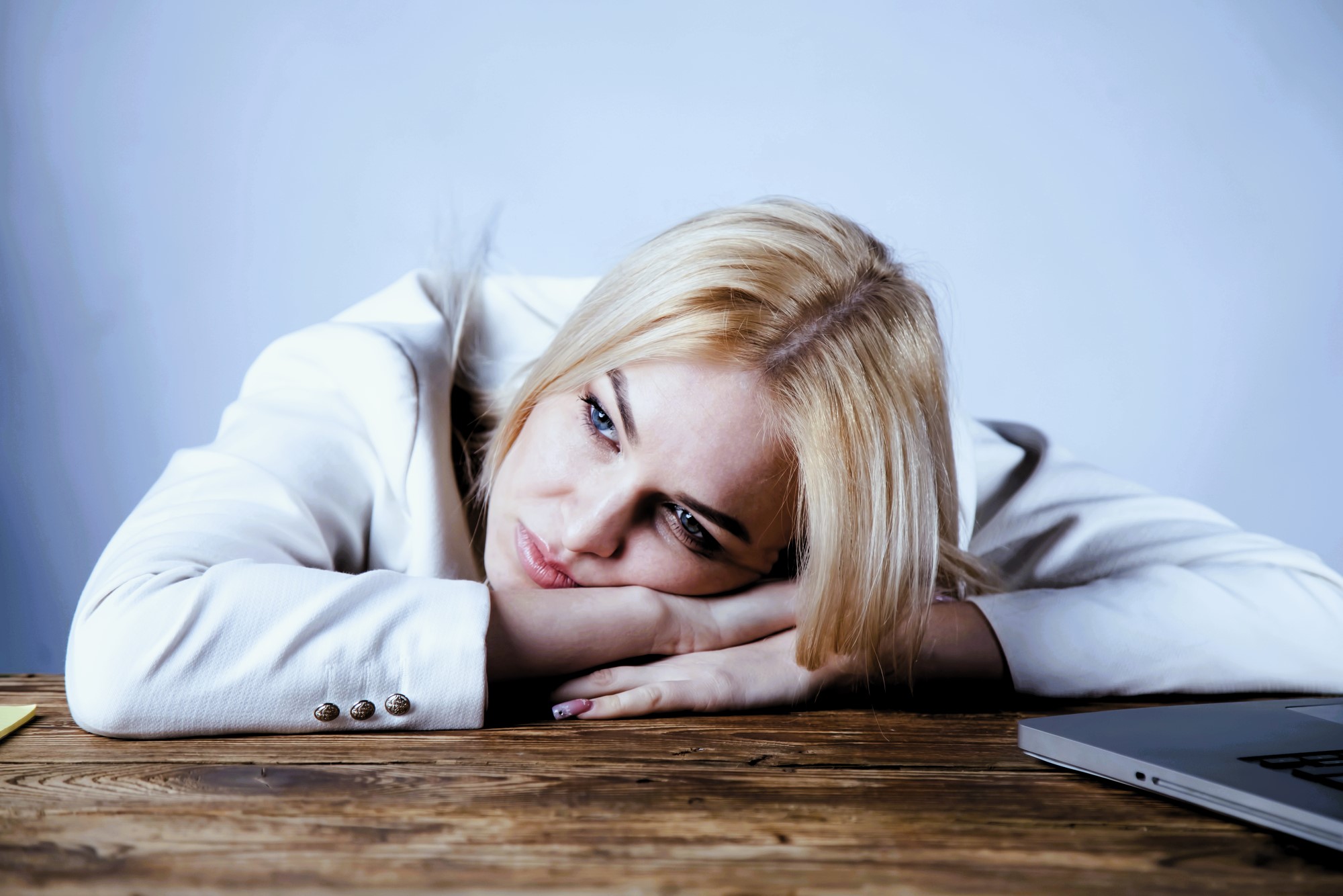 A woman with blonde hair, wearing a white blouse, rests her head on her folded arms on a wooden table. She appears thoughtful or tired, with a laptop partially visible nearby against a plain background.