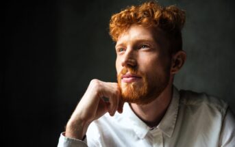 A man with curly red hair and a beard is wearing a white shirt. He is gazing thoughtfully into the distance, with one hand resting on his chin. The background is dark, creating a contrast that highlights his face.