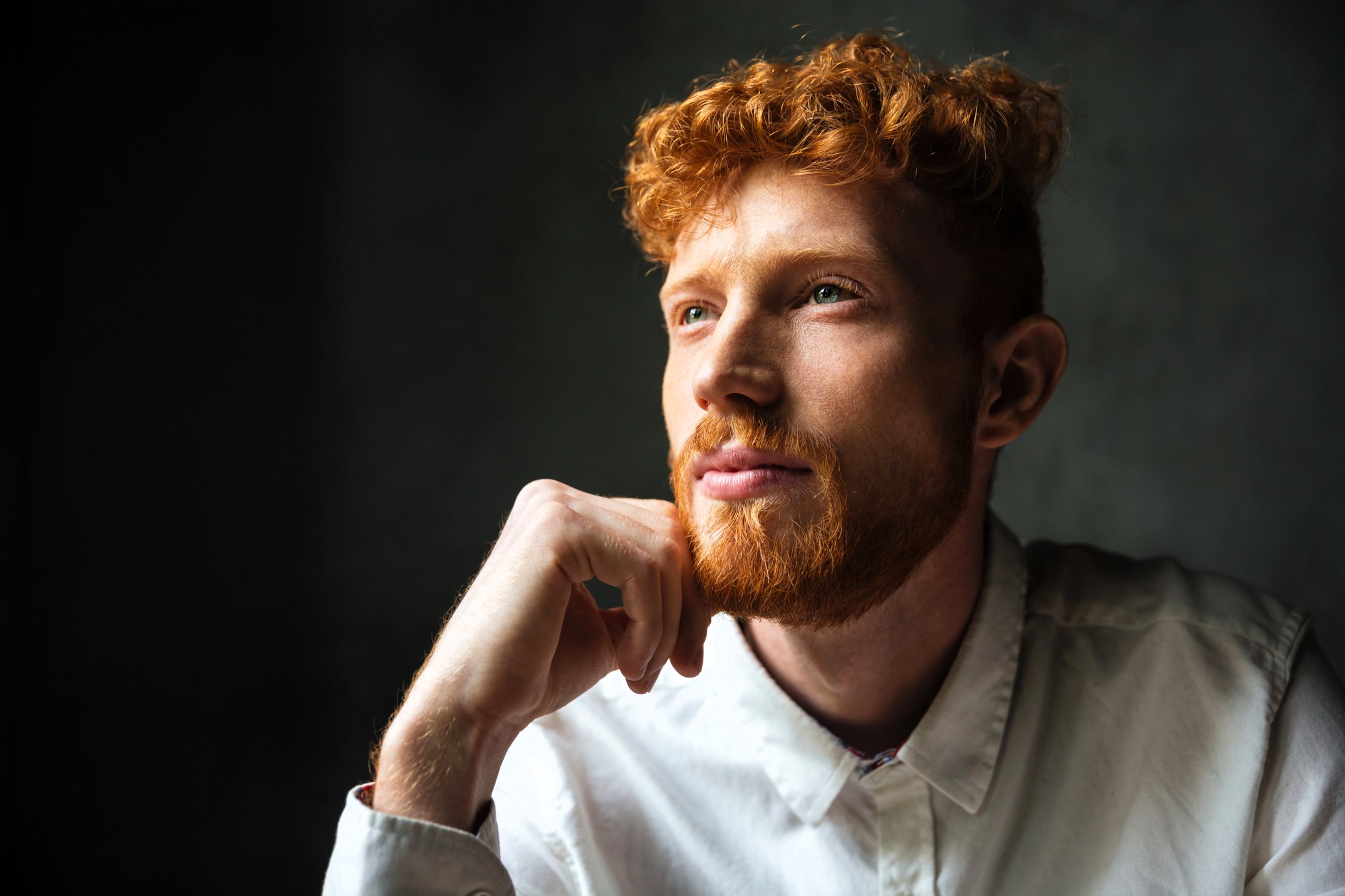 A man with curly red hair and a beard is wearing a white shirt. He is gazing thoughtfully into the distance, with one hand resting on his chin. The background is dark, creating a contrast that highlights his face.