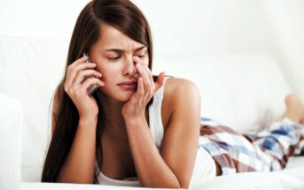 A woman with long brown hair lies on a white couch, wearing a white tank top and plaid pants. She holds a phone to her ear with one hand and touches her face with the other, appearing upset or deep in thought.