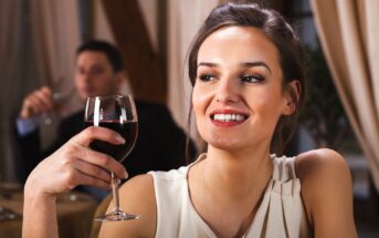 A woman in a white top, smiling and holding a glass of red wine, sits in a restaurant. A man in the background is also holding a glass. The setting is elegant, with draped curtains and warm lighting.