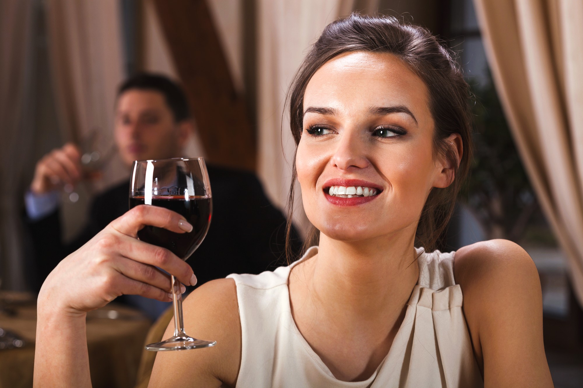 A woman in a white top, smiling and holding a glass of red wine, sits in a restaurant. A man in the background is also holding a glass. The setting is elegant, with draped curtains and warm lighting.