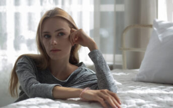 Woman with long hair, wearing a gray sweater, sits thoughtfully on a bed with a white quilt. She rests her chin on her hand and gazes out the window, with sunlight softly illuminating the room.