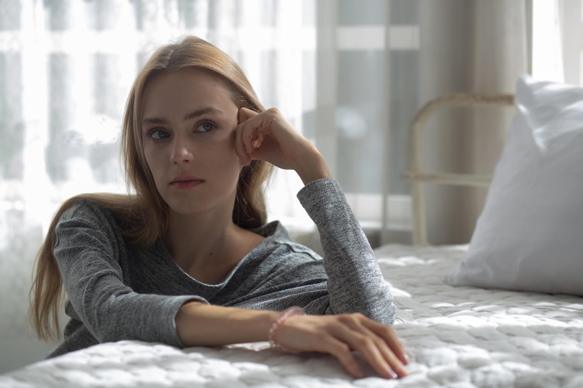 Woman with long hair, wearing a gray sweater, sits thoughtfully on a bed with a white quilt. She rests her chin on her hand and gazes out the window, with sunlight softly illuminating the room.