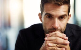 A man with short brown hair and a beard gazes intensely at the camera, resting his hands under his chin. He wears a dark jacket, and the background is softly blurred, creating a focused portrait.