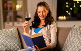 A woman with long brown hair sits on a couch, smiling while reading a book. She is holding a glass of red wine. The setting appears cozy, with throw pillows and blurred lights in the background.