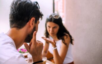 A man and a woman are sitting across from each other in a cafe, engaged in an animated conversation. The man gestures with his hand, while the woman appears to be making a point. Both are wearing sunglasses on their heads.