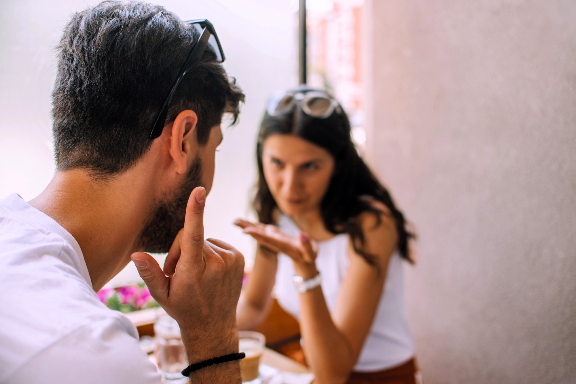 A man and a woman are sitting across from each other in a cafe, engaged in an animated conversation. The man gestures with his hand, while the woman appears to be making a point. Both are wearing sunglasses on their heads.