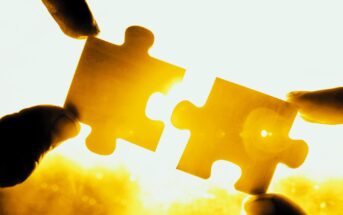 Two hands holding two puzzle pieces against a bright yellow light, with the pieces fitting together perfectly. The background is warmly lit, emphasizing the symbolic connection and completion.