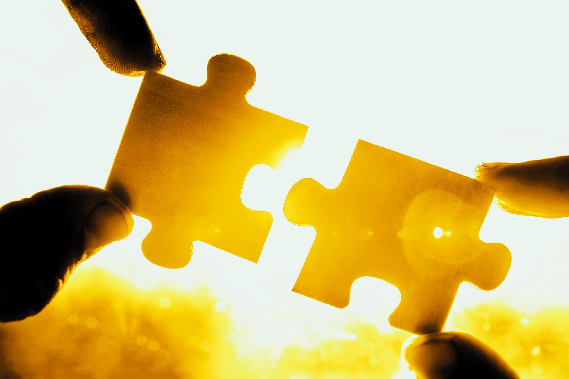 Two hands holding two puzzle pieces against a bright yellow light, with the pieces fitting together perfectly. The background is warmly lit, emphasizing the symbolic connection and completion.