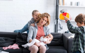 A woman sits on a couch holding a baby, while a young girl playfully climbs on her back. A boy in the foreground holds a colorful toy. The room is bright with shelves and a teddy bear visible in the background.