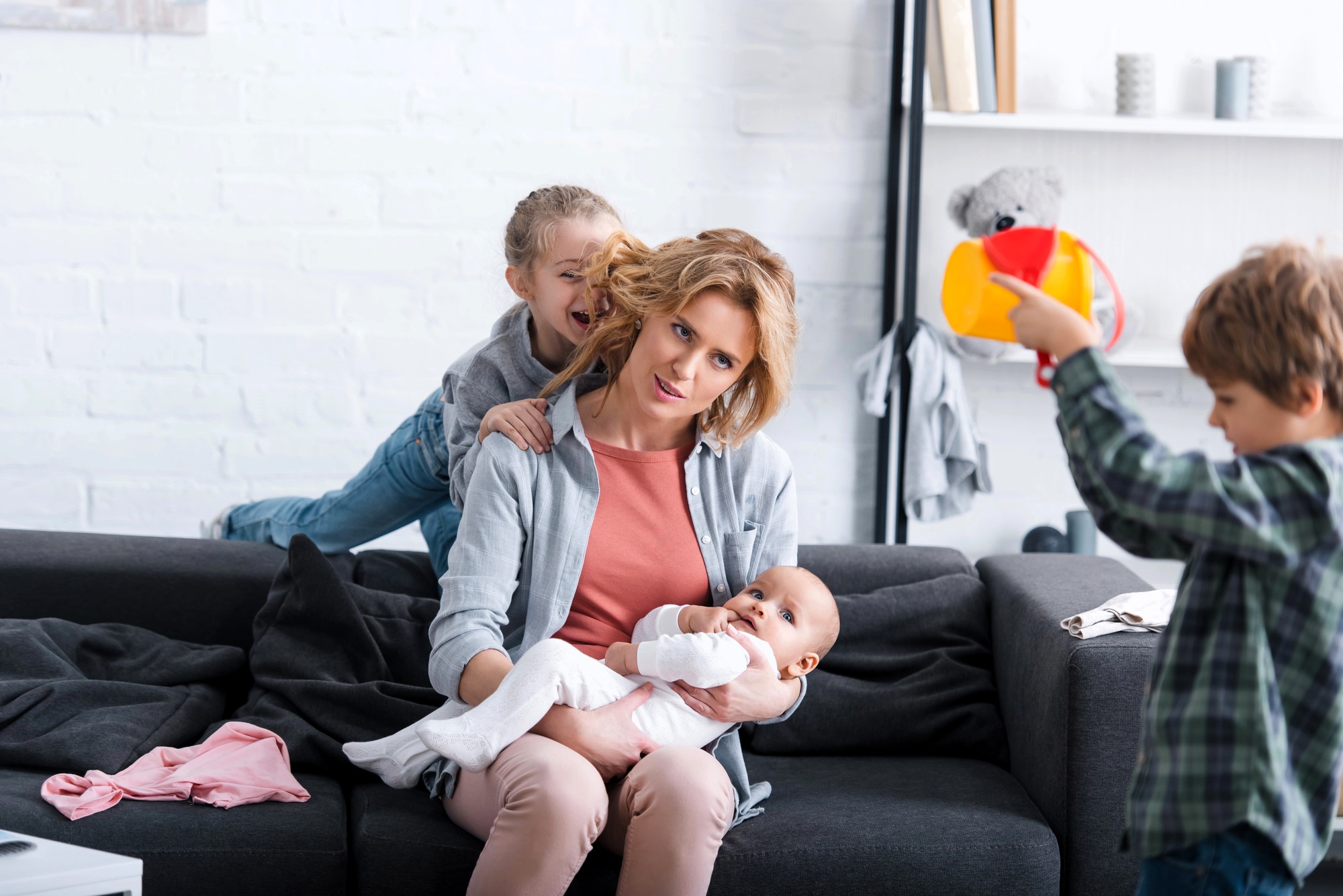 A woman sits on a couch holding a baby, while a young girl playfully climbs on her back. A boy in the foreground holds a colorful toy. The room is bright with shelves and a teddy bear visible in the background.