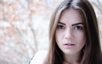 A young woman with long brown hair and expressive eyes looks towards the camera. She is wearing a light-colored sweater. The background is softly blurred with warm-toned foliage, suggesting an autumn setting.