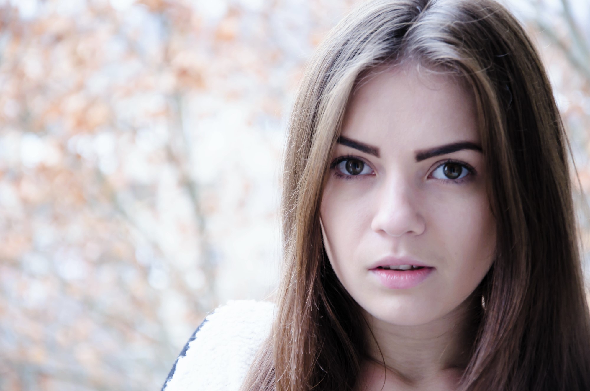 A young woman with long brown hair and expressive eyes looks towards the camera. She is wearing a light-colored sweater. The background is softly blurred with warm-toned foliage, suggesting an autumn setting.