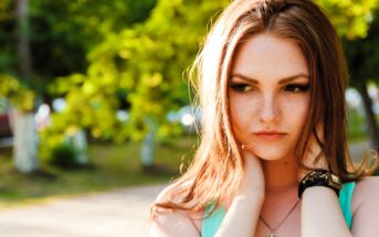 A person with long auburn hair stands outdoors, with hands near their neck. They appear contemplative, and wear a turquoise top and a bracelet. The background features green blurred foliage and sunlight.