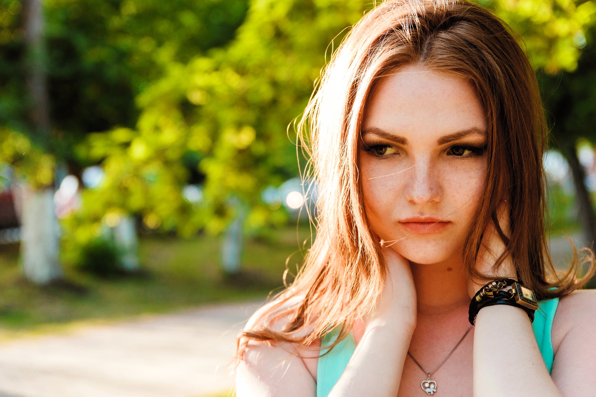 A person with long auburn hair stands outdoors, with hands near their neck. They appear contemplative, and wear a turquoise top and a bracelet. The background features green blurred foliage and sunlight.