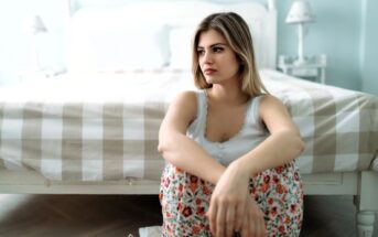 A woman with long hair sits on the floor in front of a bed. She is wearing a white tank top and floral pants. The room has a soft, muted color palette with a checkered duvet on the bed and lamps on the bedside tables.