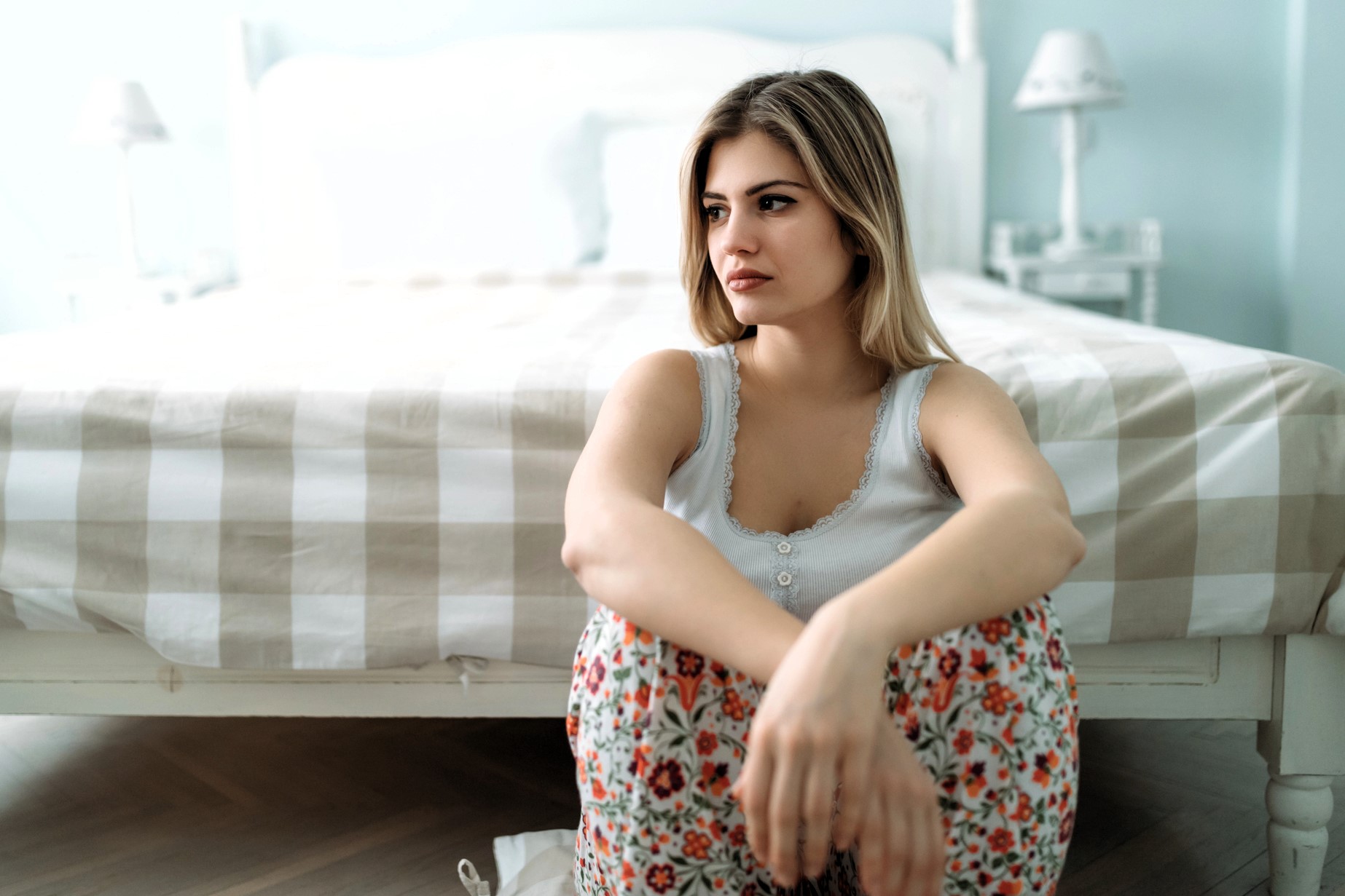 A woman with long hair sits on the floor in front of a bed. She is wearing a white tank top and floral pants. The room has a soft, muted color palette with a checkered duvet on the bed and lamps on the bedside tables.