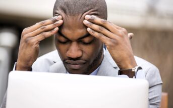 A man in a suit sits in front of a laptop, resting his forehead in his hands. His eyes are closed, and he appears deep in thought or stressed. The background is blurred and neutral.