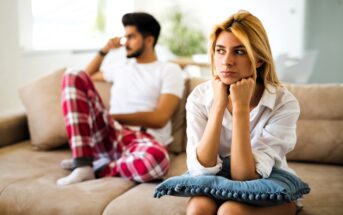 A woman with blonde hair sits on a couch, looking thoughtful and resting her chin on her hands with a blue pillow on her lap. A man in a white shirt and plaid pants sits behind her, looking away, appearing pensive.