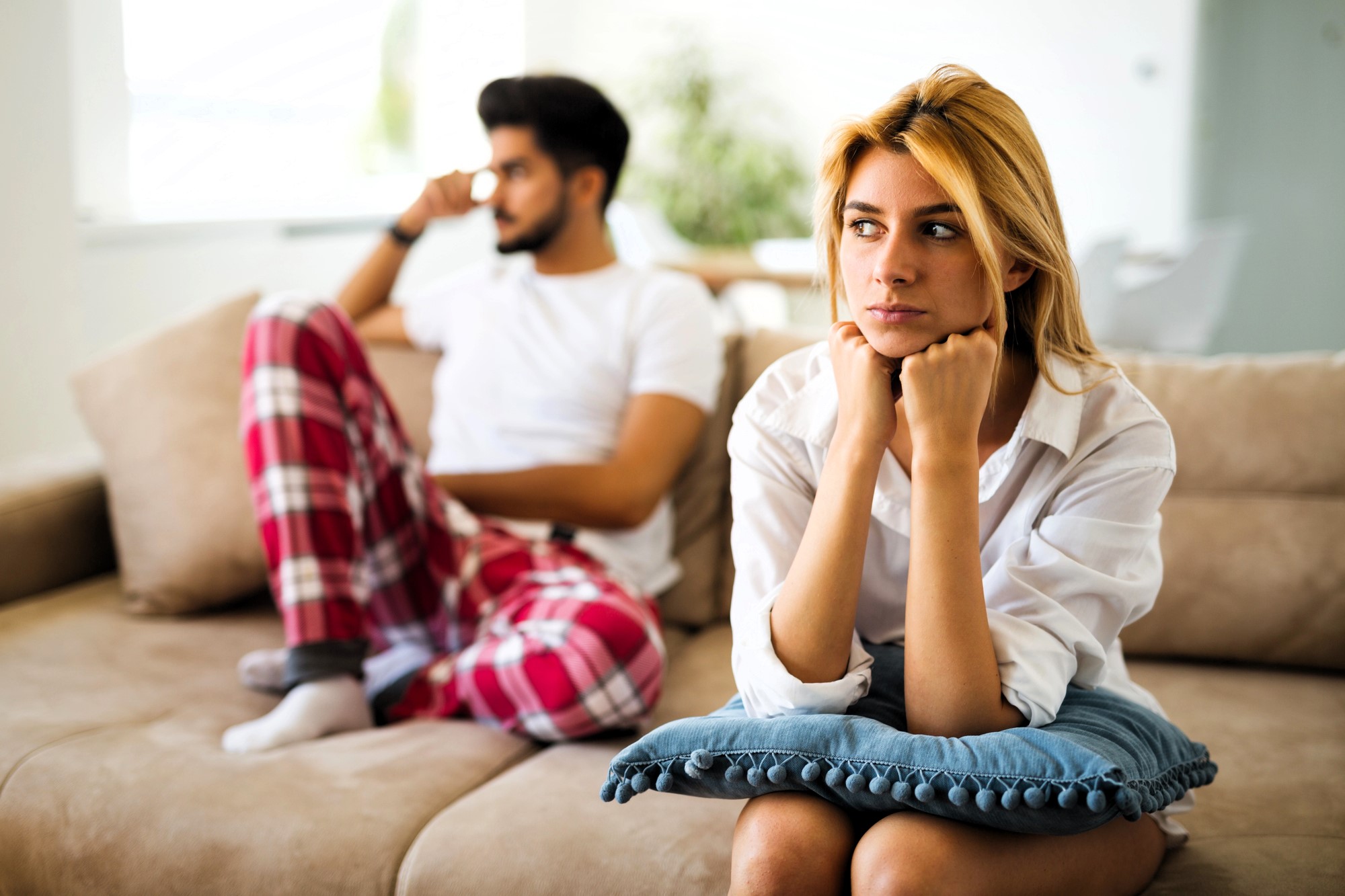 A woman with blonde hair sits on a couch, looking thoughtful and resting her chin on her hands with a blue pillow on her lap. A man in a white shirt and plaid pants sits behind her, looking away, appearing pensive.